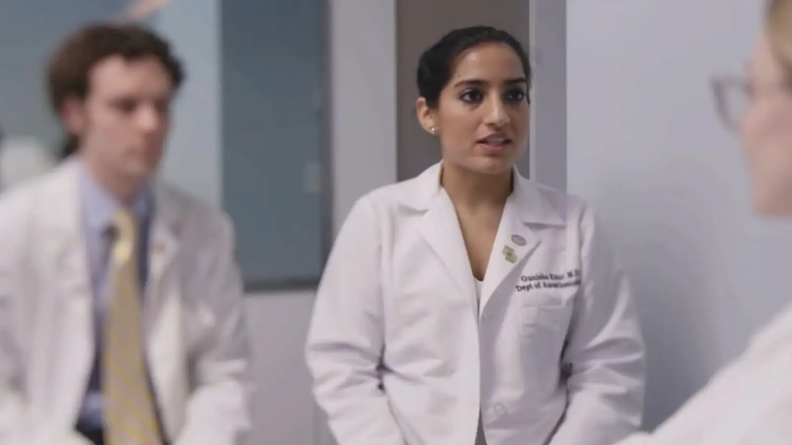 A young Indian doctor wearing a white coat smiles as she speaks to someone in front of her.