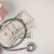 Pink piggy bank beside a calculator, U.S. currency, a stethoscope, and a red heart-shaped stress ball.