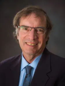 Man with glasses, salt & pepper hair wearing a suit and tie smiles at camera in professional headshot