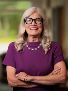 Smiling woman with silver hair, glasses and purple blouse.