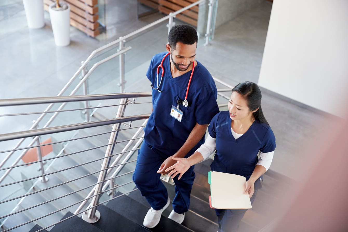 Two clinicians wearing navy blue scrubs are having a conversation and walking up a flight of stairs.