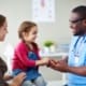 A doctor in blue scrubs holds hand of young girl during an appointment with mother looking on.