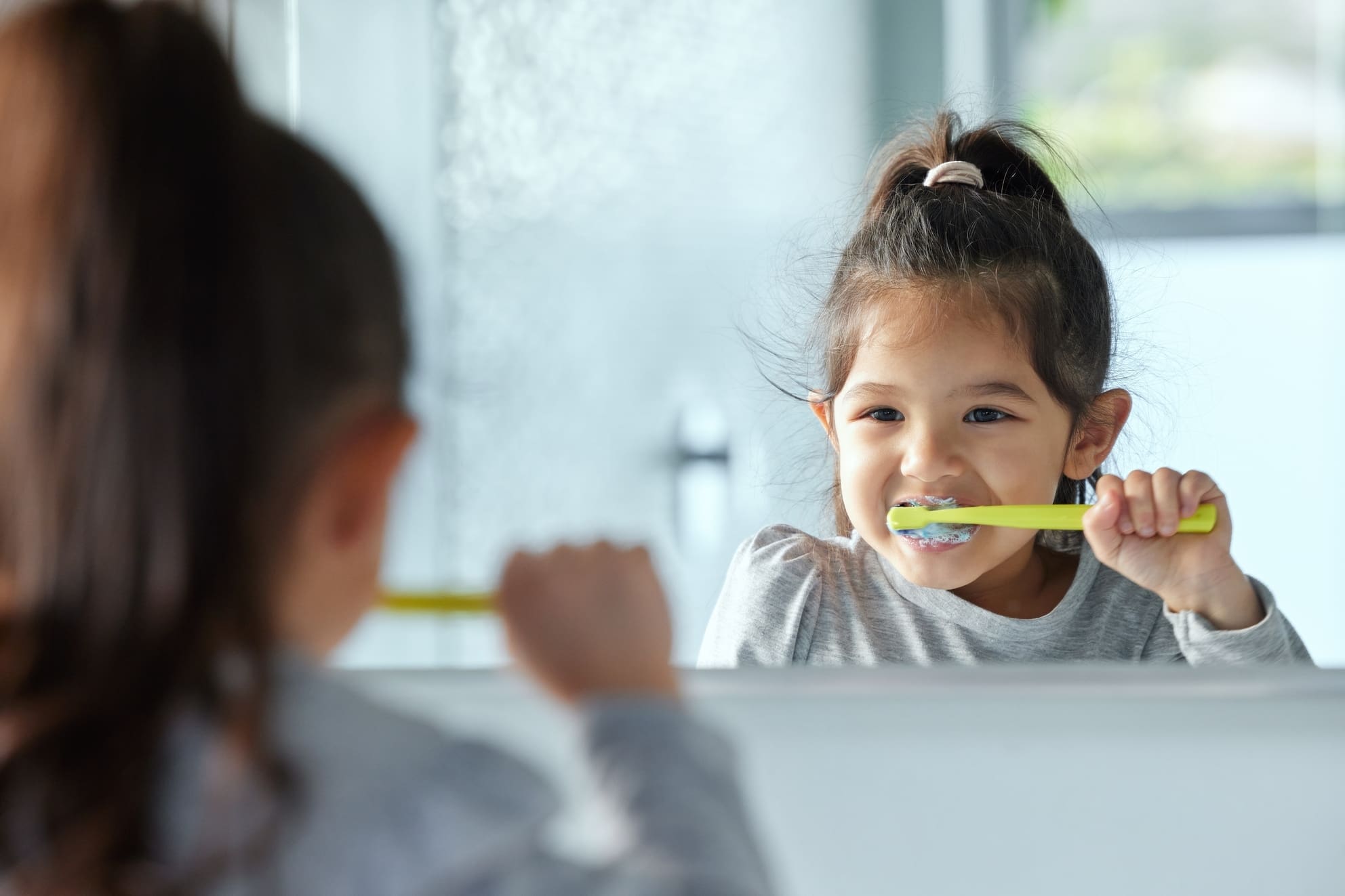 Young girl with hair in ponytail looks into a mirror and brushes her teeth.